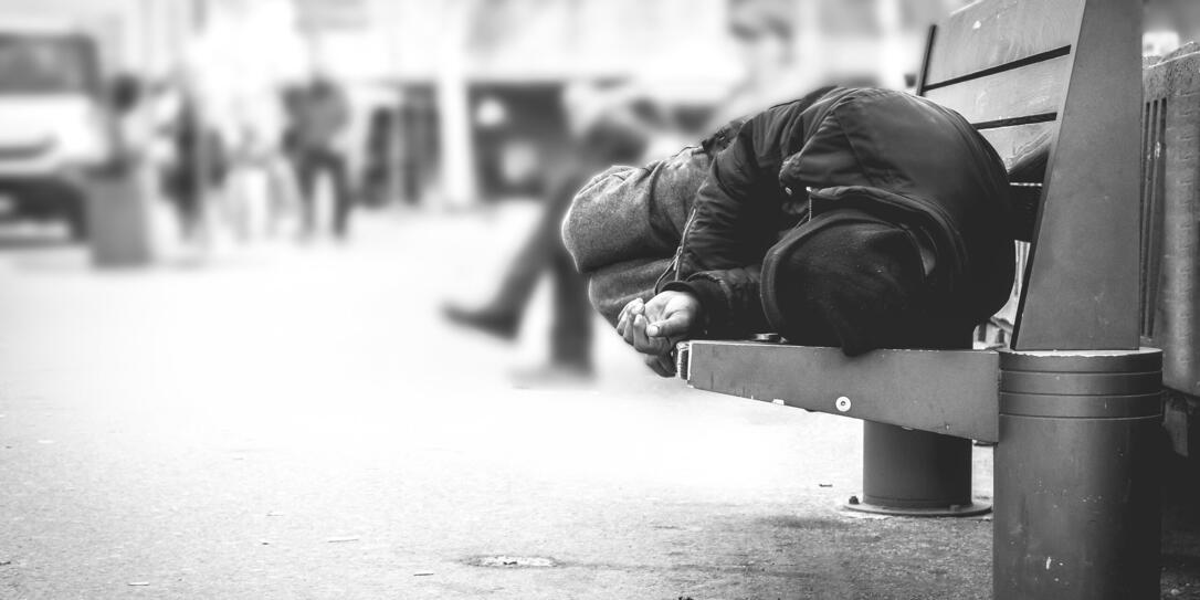 Poor homeless man or refugee sleeping on the wooden bench on the urban street in the city, social documentary concept, selective focus, black and white