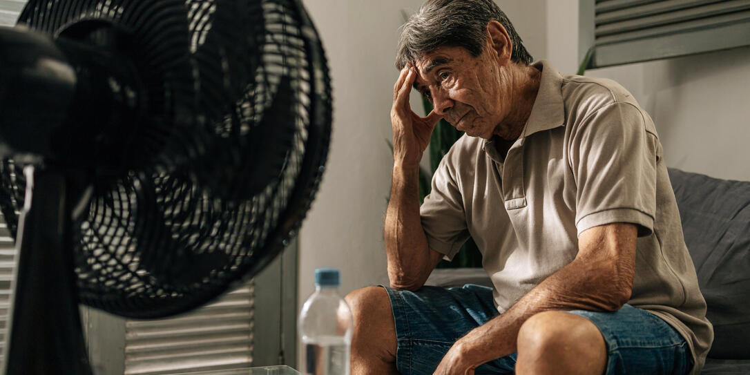 Senior man sits indoors feeling discomfort from heat while cooling himself with a fan, touching his head and resting, illustrating summer heat, fatigue, vulnerability, and everyday health concerns