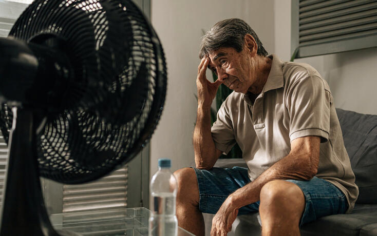 Senior man sits indoors feeling discomfort from heat while cooling himself with a fan, touching his head and resting, illustrating summer heat, fatigue, vulnerability, and everyday health concerns