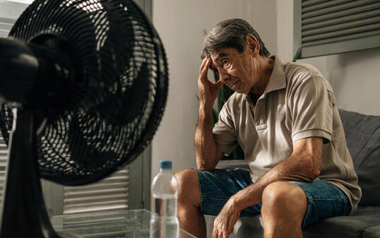 Senior man sits indoors feeling discomfort from heat while cooling himself with a fan, touching his head and resting, illustrating summer heat, fatigue, vulnerability, and everyday health concerns