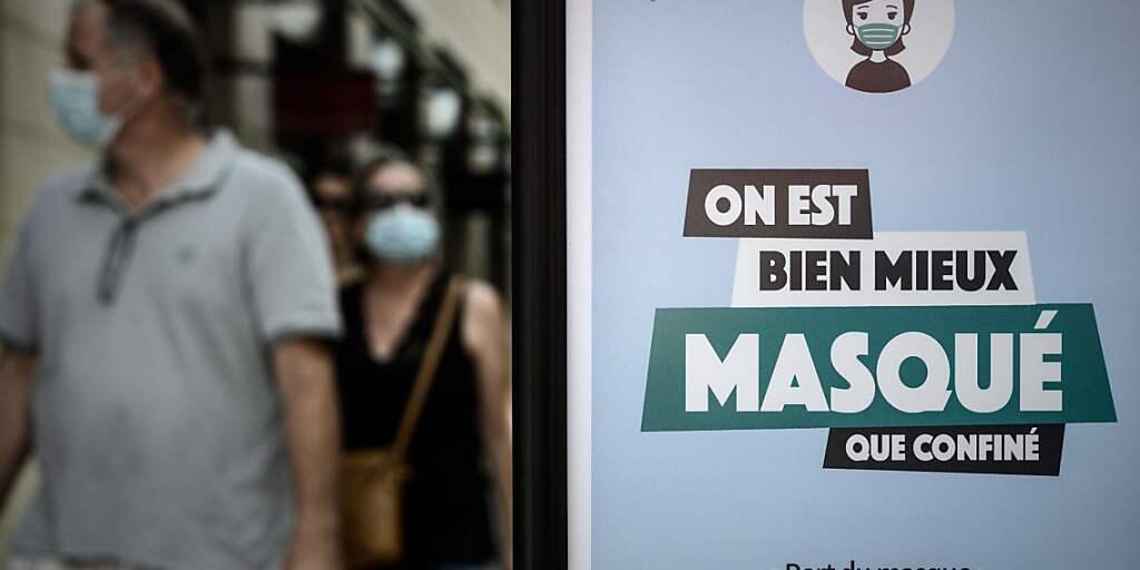 Passanten sind hinter einem Plakat mit der Aufschrift «On est bien mieux masqué que confiné» (Lieber maskiert als eingesperrt) zu sehen. Foto: Philippe Lopez/AFP/dpa